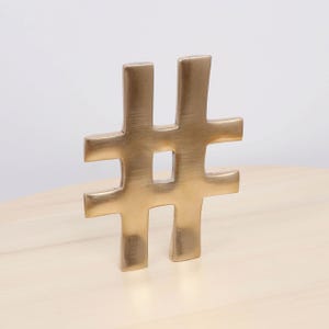 May include: A gold-coloured metal hashtag sculpture. The sculpture is a solid, three-dimensional representation of the symbol, with a square opening in the centre. It is displayed on a light-coloured wooden surface.