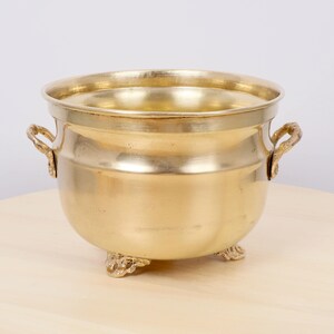 May include: A vintage brass planter. It has a rounded body, two ornate handles, and three decorative feet. The planter has a polished, reflective surface and a wide opening. The interior is visible. It sits on a light-coloured wooden surface.