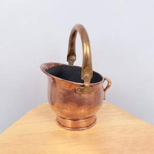 May include: A small copper bucket with a hammered finish and a brass handle. The bucket has a black interior and sits on a wooden surface.