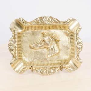 Op de afbeelding: Een goudkleurige asbak met een verhoogde Greyhound-hondenkop in het midden. De rechthoekige bak heeft een decoratieve, sierlijke rand met afgeronde hoeken en ingebouwde rustplaatsen voor sigaretten.