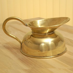 Könnte beinhalten: Eine polierte Messingkanne mit gebogenem Griff und ausgestelltem Ausguss. Die Kanne hat einen abgerundeten Boden und eine glänzende Oberfläche. Das Design ist schlicht und elegant, geeignet zum Servieren von Getränken oder als dekoratives Element. Ca. 20 cm hoch.