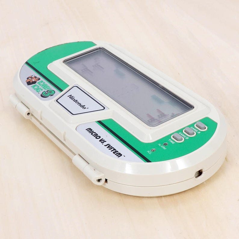 May include: A vintage Nintendo Micro Vs. System handheld game console. The device is white with green accents and features a small screen displaying a game. The console has buttons for Game A, Game B, Time, and Alarm.