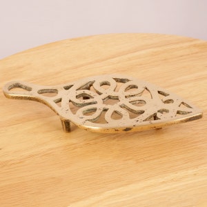 May include: A brass trivet with a decorative, intricate design. The trivet has a handle and three legs.