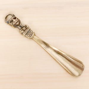 May include: A metallic gold-toned shoehorn featuring a decorative handle shaped like a cat face with pointed ears, positioned above a textured brick-patterned base. The long, smooth, curved shaft extends downward to a rounded, tapered end.
