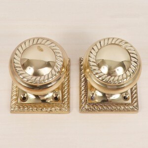 May include: Two polished brass door knobs featuring a round, smooth center and a decorative rope-textured border. Each knob is mounted on a square brass backplate with a matching rope-textured edge and two visible screw holes for installation.