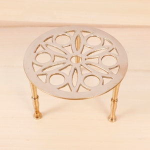 Puede incluir: Un salvamanteles de metal dorado con un diseño calado decorativo. El salvamanteles tiene una parte superior circular con un patrón floral y tres patas cortas. Está diseñado para proteger las superficies de los utensilios de cocina calientes.