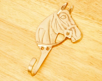 May include: A gold-colored metal horse head coat hook. The hook is shaped like a horse's head with a curved hook at the bottom.