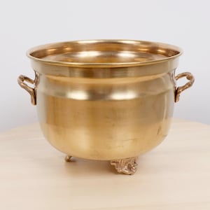 Puede incluir: Una maceta de latón pulido con un cuerpo ancho y redondeado y una superficie lisa y reflectante. El recipiente cuenta con dos pequeñas asas ornamentadas en los lados opuestos y descansa sobre tres pies decorativos esculpidos y estables.