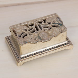 May include: A brass trinket box with an intricate floral design. The box has a raised, decorative border and a hinged lid.
