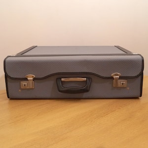 May include: A gray suitcase with black trim and a black handle. The suitcase has two silver latches on the front.