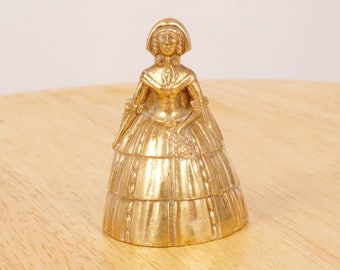 May include: A gold-colored brass bell shaped like a woman in a long dress. The bell has a detailed design and is sitting on a wooden surface.
