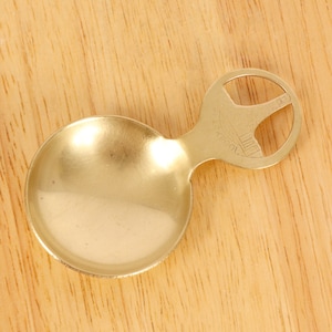 May include: A gold-colored metal teaspoon with a unique handle design. The handle has a cut-out shape and the word "TOOK" is engraved on it.