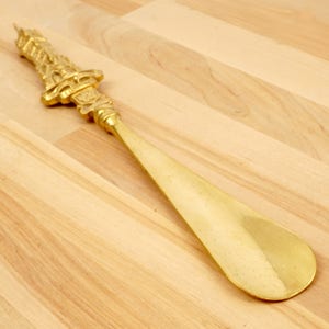 Peut inclure: Un chausse-pied en m&eacute;tal dor&eacute; avec une poign&eacute;e orn&eacute;e et d&eacute;corative. Le chausse-pied est sur une surface en bois clair.