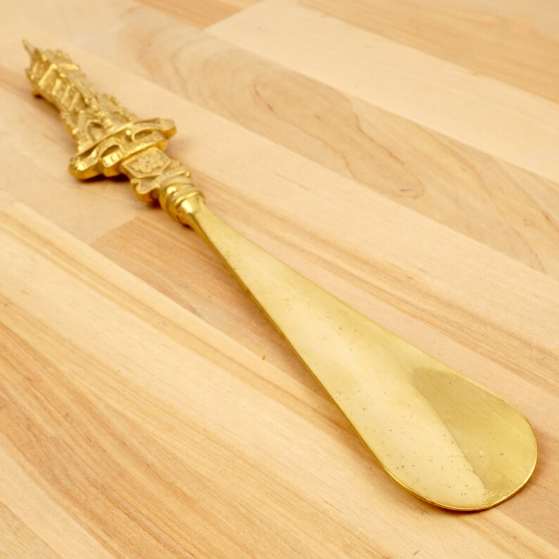 Peut inclure: Un chausse-pied en m&eacute;tal dor&eacute; avec une poign&eacute;e orn&eacute;e et d&eacute;corative. Le chausse-pied est sur une surface en bois clair.