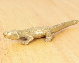 May include: A gold-colored alligator figurine with a textured surface. The figurine is posed in a walking position with its mouth closed.