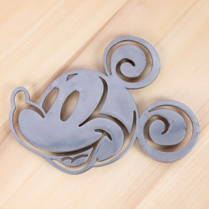 May include: A silver metal cutout of Mickey Mouse's head. The cutout is a simple design with Mickey's ears, eyes, and smile.
