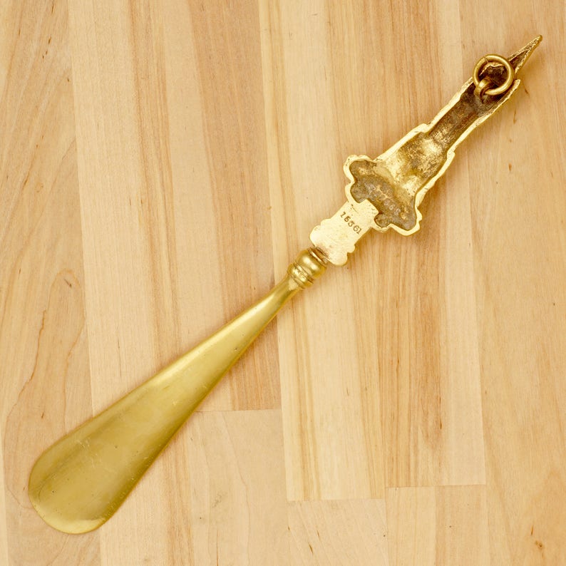 Peut inclure: Un chausse-pied en or avec une poign&eacute;e d&eacute;corative. La poign&eacute;e pr&eacute;sente un motif en relief et le num&eacute;ro "1861" est grav&eacute; sur la poign&eacute;e.