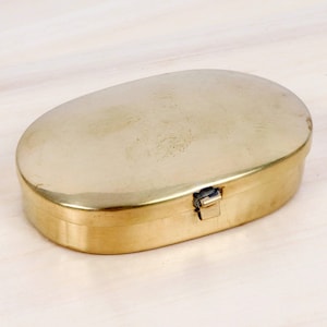 Könnte beinhalten: Eine ovale Messingdose mit einer glatten, polierten metallischen Oberfläche. Der Behälter verfügt über einen Klappdeckel, der mit einem kleinen, integrierten Metallverschlussmechanismus an der Vorderseite des Dosenkörpers sicher schließt.