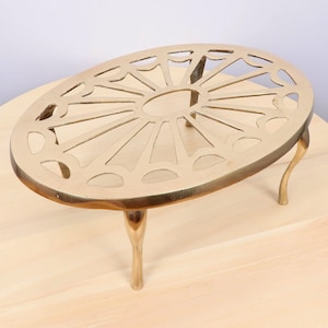 May include: A gold-colored trivet with an oval shape and a decorative, openwork design. The trivet has three curved legs for stability. The openwork pattern features a central oval surrounded by radiating lines and arched cutouts.