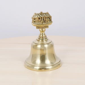 May include: A brass bell with a detailed miniature building on top. The building has a thatched roof and is inscribed with "The Hoops in Horwood Cross, North Devon". The bell is on a light wood surface.