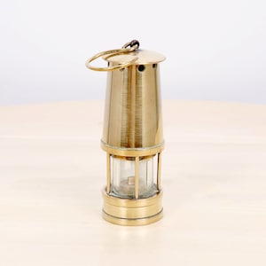 Op de afbeelding: Een vintage messing mijnwerkerslamp met een heldere glazen behuizing en een ronde handgreep. De lamp heeft een conische vorm met een afgeronde bovenkant en een voet. Het messing heeft een gepolijste afwerking.