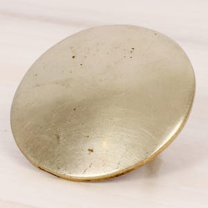 Può includere: Oggetto metallico ovale e usurato, con superficie spazzolata. Il bordo è leggermente rialzato. Sembra essere in ottone o metallo simile. Diametro di circa 10 cm.
