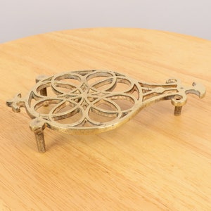 Puede incluir: Un salvamanteles de metal dorado con un diseño decorativo intrincado. El salvamanteles tiene un asa y tres patas.