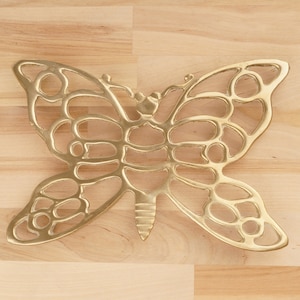 May include: A gold metal butterfly-shaped trivet with a decorative cutout design. The trivet is on a light wood surface.