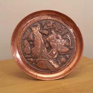Puede incluir: Un plato de cobre con un diseño en relieve de personas con vestimenta tradicional. El plato es redondo y tiene un borde elevado. El diseño es intrincado y detallado, y el plato está en buen estado.