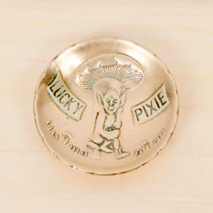 Puede incluir: Un plato redondo y metálico con un diseño en relieve que presenta un duende sentado bajo una seta. Las palabras "LUCKY" y "PIXIE" están inscritas en pancartas. El plato tiene un borde festoneado y está sobre un fondo de madera clara.