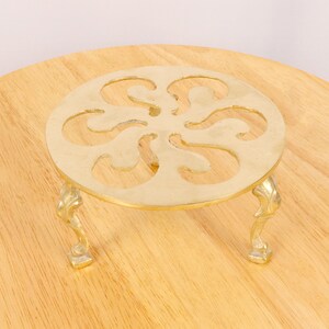 May include: A gold-coloured metal trivet with a decorative, circular design and three legs. The trivet is on a wooden surface.