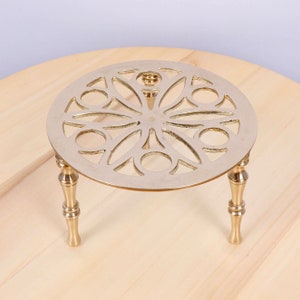 Puede incluir: Un soporte decorativo de metal dorado con una parte superior redonda y tres patas. La parte superior tiene un diseño circular calado.