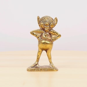 Könnte beinhalten: Eine goldfarbene Messingfigur eines schelmischen Gnoms mit einem breiten Grinsen und großen Ohren. Der Gnom steht mit den Händen in den Hüften und trägt einen einfachen, abgerundeten Kragen. Die Figur ist etwa 12,5 cm hoch und steht auf einem kleinen, rechteckigen Sockel.