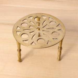 Puede incluir: Un salvamanteles redondo de latón con un diseño calado decorativo. El salvamanteles tiene tres patas cortas y una serie de formas recortadas. Está diseñado para proteger las superficies de la cocina caliente.