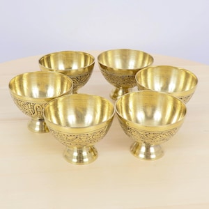 May include: Six gold-coloured metal bowls with ornate, etched designs. The bowls have a pedestal base and a rounded bowl shape. The bowls are arranged on a light-coloured wooden surface.
