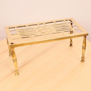 May include: A brass pot stand with a decorative geometric design and four legs. The stand is made of polished brass and has a rectangular shape.