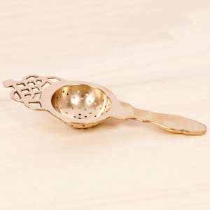 May include: A gold-toned metal tea strainer featuring a perforated bowl and a decorative handle with an openwork knot design. The strainer has a smooth, polished finish and a long, flat handle extending from the circular, bowl-shaped filter.