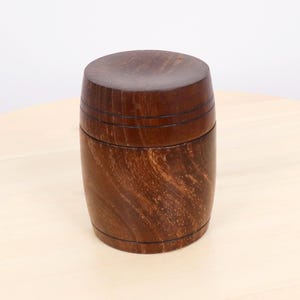 Puede incluir: Un recipiente artesanal de madera en forma de barril con tapa. El recipiente es de madera marrón oscuro con vetas visibles y ranuras horizontales. La tapa tiene una forma sólida y redondeada. Parece ser un objeto de almacenamiento.