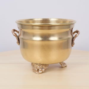 Può includere: Un calderone in metallo color oro con due manici ornati e tre piedini decorativi. Il calderone ha un corpo arrotondato e un bordo largo. I manici e i piedini hanno un design dettagliato e strutturato.