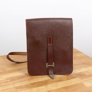 May include: A rectangular, brown leather shoulder bag with a textured surface. The bag features a long, thin strap and a vertical strap with a metal buckle closure. The bag is sitting on a wooden surface.
