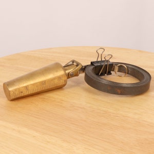 May include: A brass measuring tape with a graduated scale and a metal weight attached to the end. The tape is rolled up and secured with a pair of binder clips.
