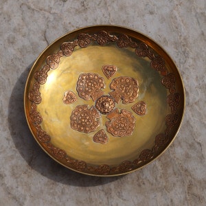 May include: A round, gold-colored metal serving dish with intricate copper-colored designs. The dish has a raised center and a decorative border.