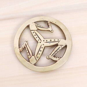 Puede incluir: Un salvamanteles circular de latón con el símbolo del Triskelion, un diseño de tres piernas dobladas que irradian desde un punto central. El salvamanteles tiene un borde decorativo.