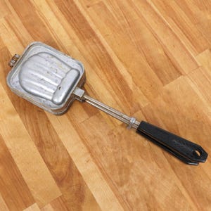 May include: A silver-coloured, handheld sandwich maker with a black handle. The maker has a ridged, textured surface and is designed to create a sandwich with a scalloped edge.