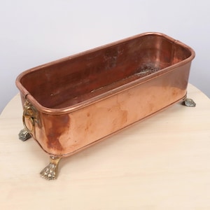 May include: A rectangular copper planter with a polished finish, featuring four ornate metal claw feet and two lion head handles on the sides, each holding a circular ring. The interior is deep and open, showing a smooth metallic surface.
