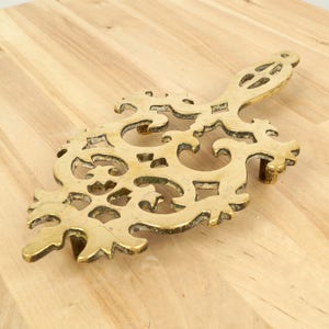 Peut inclure: Un dessous de plat en métal doré avec un design complexe et orné. Le dessous de plat a une poignée d'un côté et est parfait pour protéger les surfaces des plats chauds.