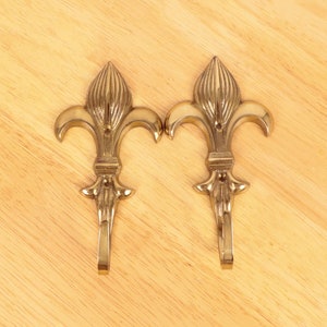 Peut inclure: Deux crochets en forme de fleur de lys de couleur laiton. Chaque crochet présente un motif floral stylisé avec un élément vertical central et trois pétales incurvés. Les crochets sont posés sur une surface en bois clair.