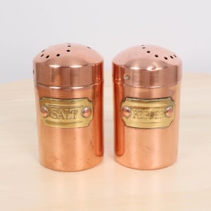 May include: Two copper salt and pepper shakers with brass labels. The salt shaker has "SALT" on the label, and the pepper shaker has "PEPPER" on its label. Both shakers have a cylindrical shape with a rounded top and small holes.
