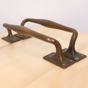 Op de afbeelding: Twee antieke messing deurgrepen met een gebogen ontwerp en een vierkante basis. De grepen bevinden zich op een licht houten oppervlak.