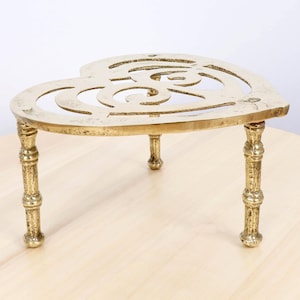 Puede incluir: Un salvamanteles de latón en forma de corazón con tres patas ornamentadas. El salvamanteles tiene un diseño calado decorativo en el centro. El latón tiene un color dorado cálido y está diseñado para proteger las superficies de los objetos calientes.